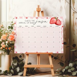 May include: A pink and white baby shower calendar on a wooden easel. The calendar has a strawberry design and the text "Guess the Arrival of Baby Jensen". The calendar is surrounded by flowers.