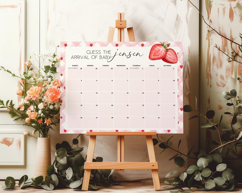 May include: A pink and white baby shower calendar on a wooden easel. The calendar has a strawberry design and the text "Guess the Arrival of Baby Jensen". The calendar is surrounded by flowers.