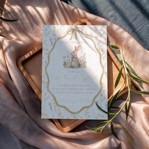 May include: An Easter-themed invitation card with a bunny illustration. The card features a decorative border and text that reads "Hop-on-over for an Easter Special - Egg Hunt". The card is on a wooden tray and a peach-colored fabric.