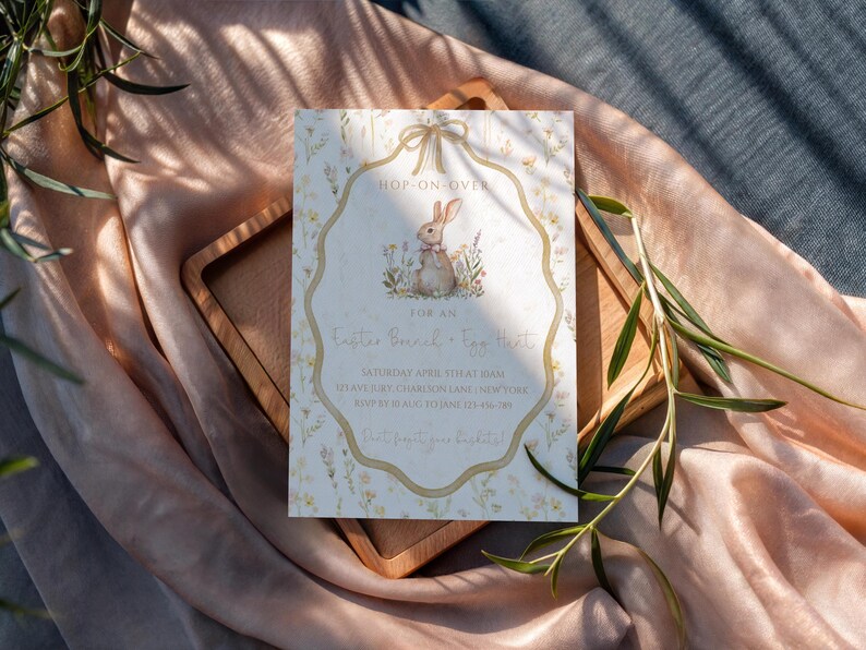 May include: An Easter-themed invitation card with a bunny illustration. The card features a decorative border and text that reads "Hop-on-over for an Easter Special - Egg Hunt". The card is on a wooden tray and a peach-colored fabric.