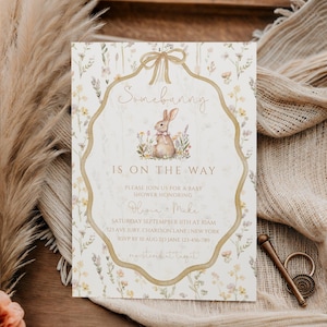 May include: Baby shower invitation with a floral design and a bunny illustration. The text reads "Somebunny is on the way" and includes event details. The invitation is on a wooden tray with dried plants and fabric.