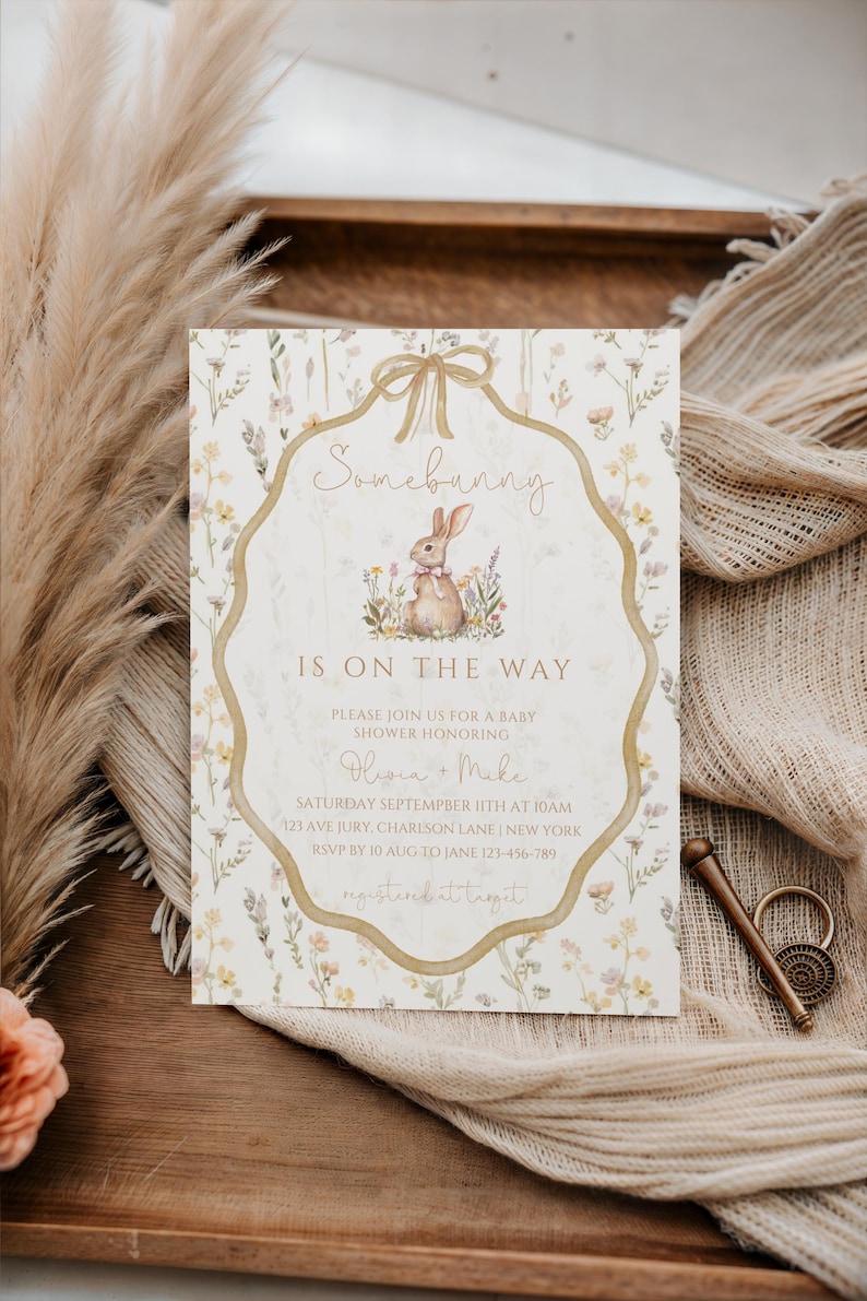 May include: Baby shower invitation with a floral design and a bunny illustration. The text reads "Somebunny is on the way" and includes event details. The invitation is on a wooden tray with dried plants and fabric.