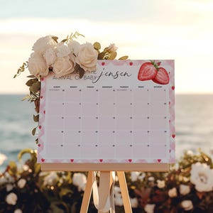 May include: A baby shower calendar on a wooden easel, decorated with white roses and greenery. The calendar has a pink border with hearts and strawberries, and the text "Arrival of Baby Jensen".