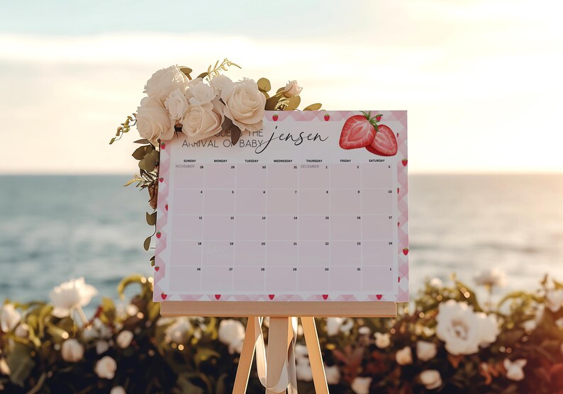 May include: A baby shower calendar on a wooden easel, decorated with white roses and greenery. The calendar has a pink border with hearts and strawberries, and the text "Arrival of Baby Jensen".