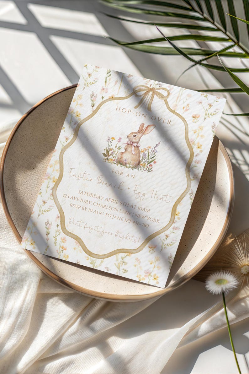 May include: An Easter brunch and egg hunt invitation card with a watercolor bunny illustration. The invitation is on a cream-colored background with floral accents and the text "Hop-On-Over". The card is on a beige plate.