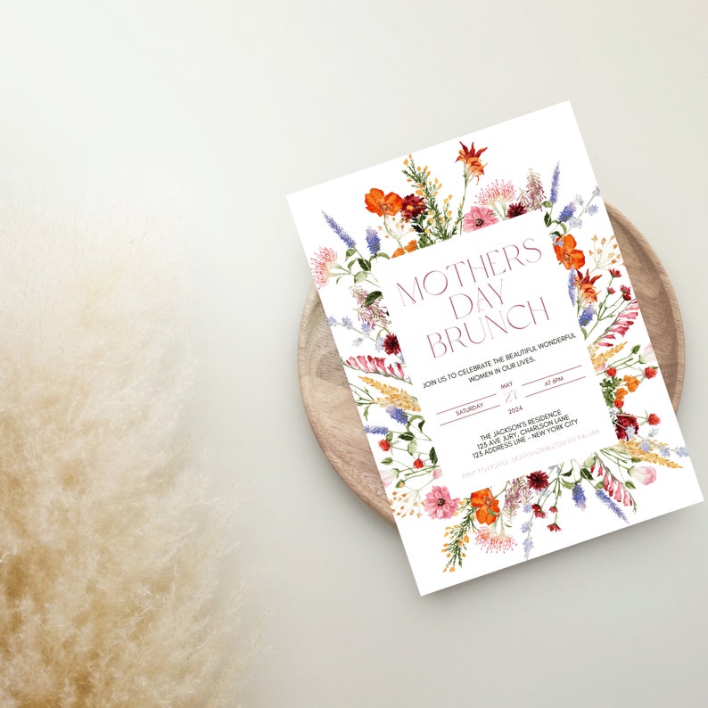 Pu&ograve; includere: Un invito per il brunch della festa della mamma con bordo floreale. L'invito presenta il testo "Mothers Day Brunch" in un carattere bordeaux, circondato da una composizione floreale ad acquerello di fiori arancioni, rosa e viola. L'invito &egrave; su un piatto di legno.