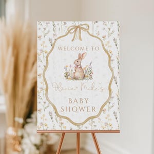 Può includere: Un cartello di benvenuto per il baby shower con un'illustrazione ad acquerello di un coniglietto con un fiocco rosa, circondato da fiori. Il cartello recita "Benvenuti al baby shower di Olivia + Mike" in una scrittura elegante su uno sfondo floreale.