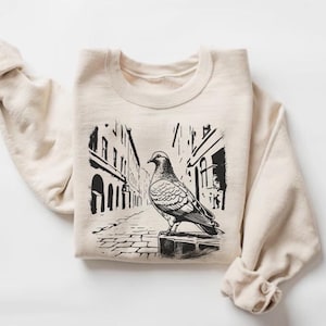 May include: A beige sweatshirt with a black and white illustration of a pigeon standing in a city street. The illustration is in a vintage style.