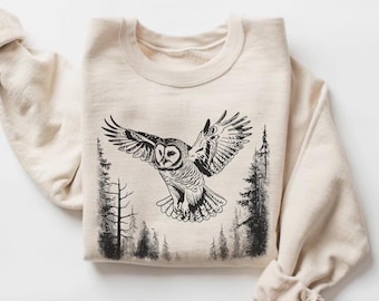 Può includere: Una felpa beige con un'illustrazione in bianco e nero di un gufo che vola attraverso un bosco. Il gufo è al centro dell'immagine e gli alberi sono su entrambi i lati.