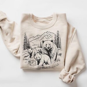 May include: A beige sweatshirt with a black and white illustration of three bears in a mountain landscape. The bears are standing in a stream of water.