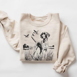 Può includere: Una felpa beige con un'illustrazione in bianco e nero di un cane che punta a tre uccelli in volo. Il cane è in piedi nell'erba alta.