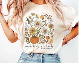 May include: Cream-colored t-shirt featuring a floral and pumpkin graphic. The design includes daisies, leaves, and pumpkins in shades of yellow, orange, and green. The text "in all things, give thanks" is printed below the graphic.