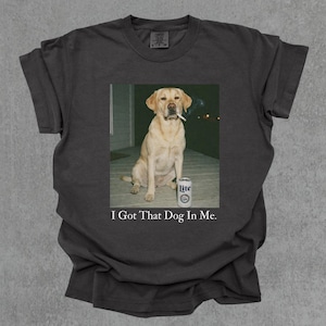 Peut inclure: T-shirt gris foncé avec un imprimé d'un Labrador jaune fumant une cigarette, avec une canette de bière Lite. Le texte "I Got That Dog In Me" est imprimé en dessous.