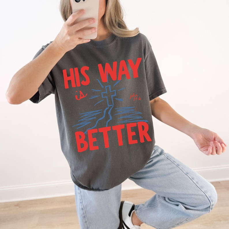 Woman wearing a Christian graphic t-shirt with red text “His Way Is Better” and a blue cross illustration with John 14:6.