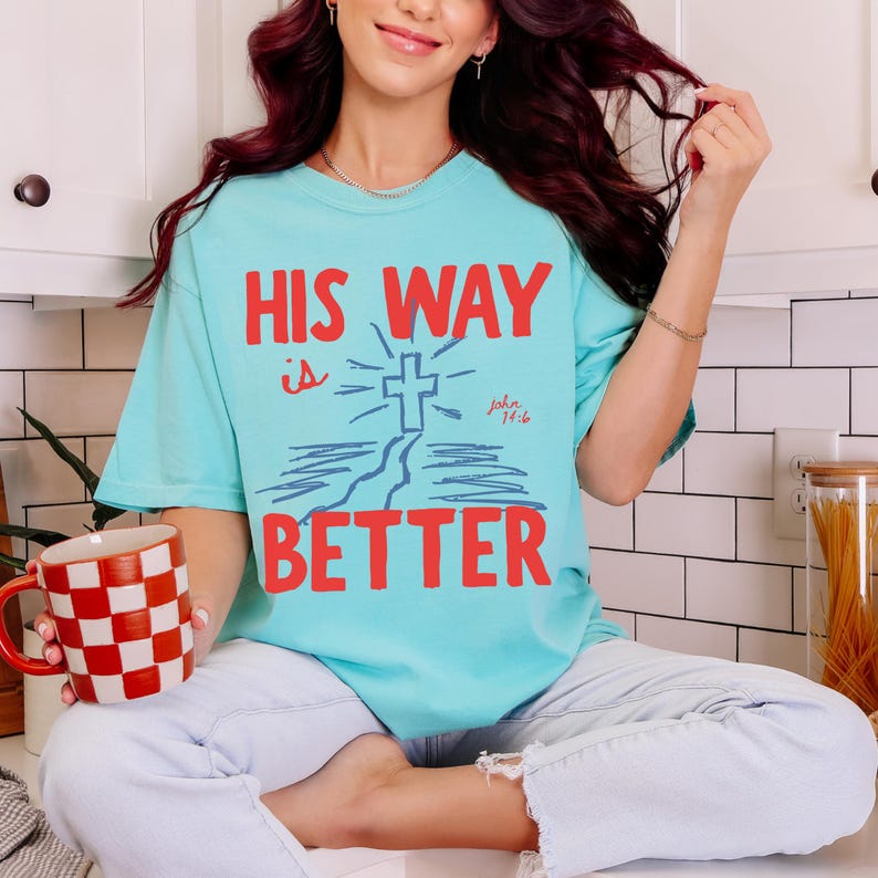 Woman wearing a Christian graphic t-shirt with red text “His Way Is Better” and a blue cross illustration with John 14:6.