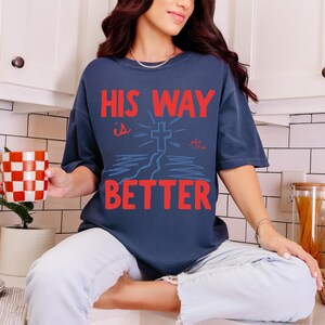 Woman wearing a Christian graphic t-shirt with red text “His Way Is Better” and a blue cross illustration with John 14:6.