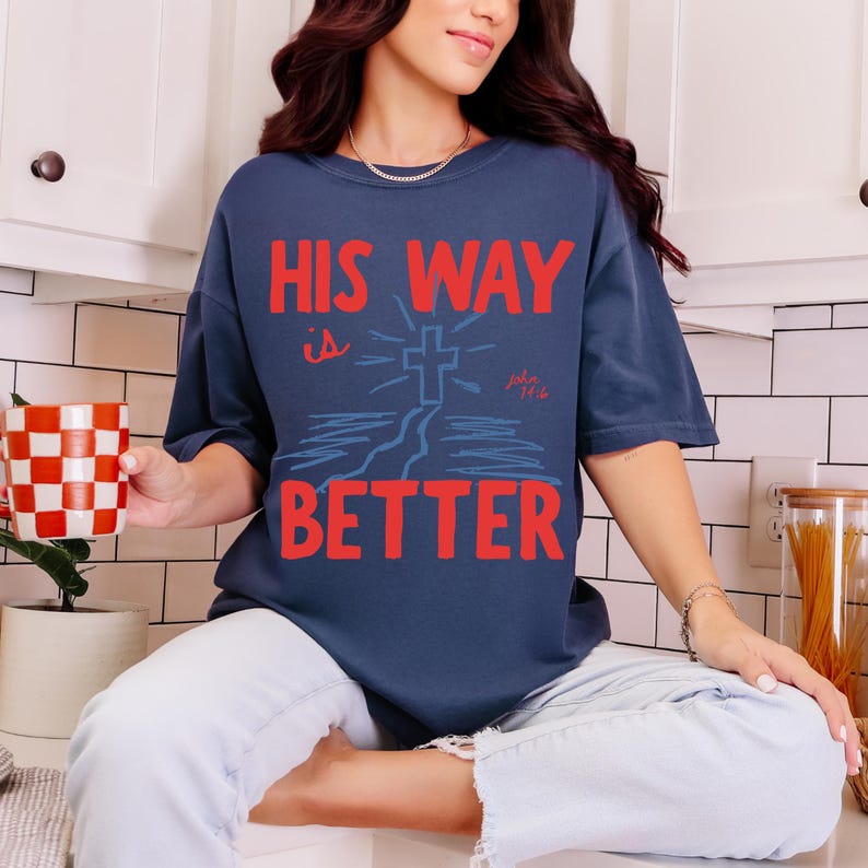 Woman wearing a Christian graphic t-shirt with red text “His Way Is Better” and a blue cross illustration with John 14:6.