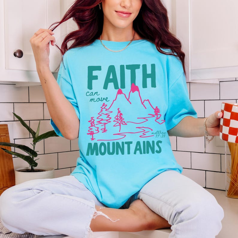Woman wearing a Christian graphic t-shirt with colorful “Faith can move Mountains” text, pink mountain illustration, and Matthew 17:20 Bible verse.
