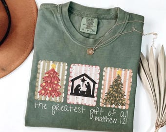 以下が含まれることがあります： オリーブグリーンのTシャツ。クリスマスツリー、降誕シーン、2つ目のクリスマスツリーのグラフィックが特徴です。「the greatest gift of all matthew 1:21」のテキストが下に印刷されています。小さなペンダントが付いたシルバーのネックレスがシャツの上にあります。