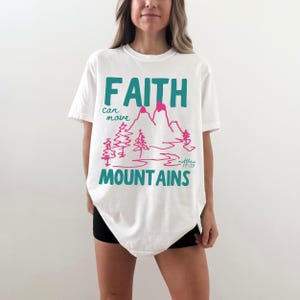 Woman wearing a Christian graphic t-shirt with colorful “Faith can move Mountains” text, pink mountain illustration, and Matthew 17:20 Bible verse.