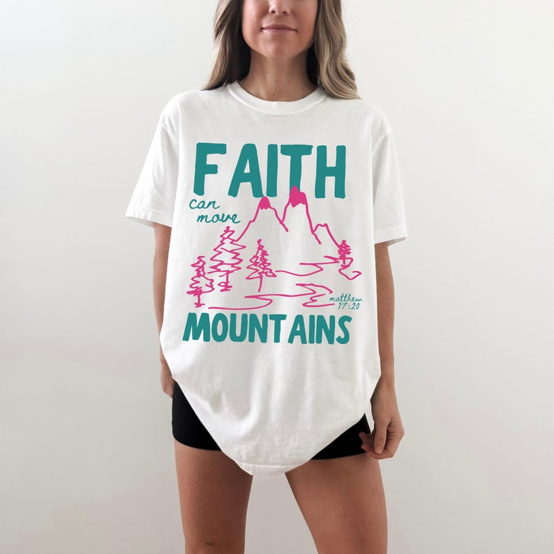 Woman wearing a Christian graphic t-shirt with colorful “Faith can move Mountains” text, pink mountain illustration, and Matthew 17:20 Bible verse.