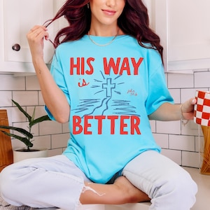 Woman wearing a Christian graphic t-shirt with red text “His Way Is Better” and a blue cross illustration with John 14:6.