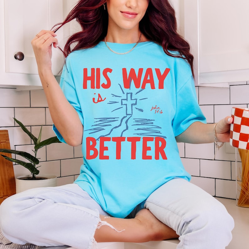 Woman wearing a Christian graphic t-shirt with red text “His Way Is Better” and a blue cross illustration with John 14:6.