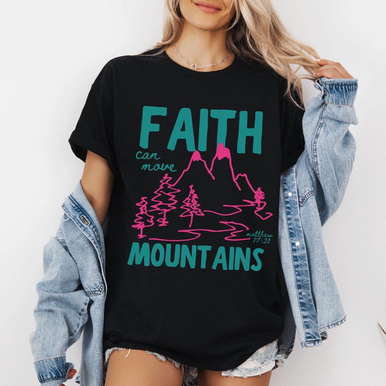 Woman wearing a Christian graphic t-shirt with colorful “Faith can move Mountains” text, pink mountain illustration, and Matthew 17:20 Bible verse.
