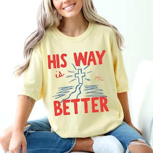 Woman wearing a Christian graphic t-shirt with red text “His Way Is Better” and a blue cross illustration with John 14:6.