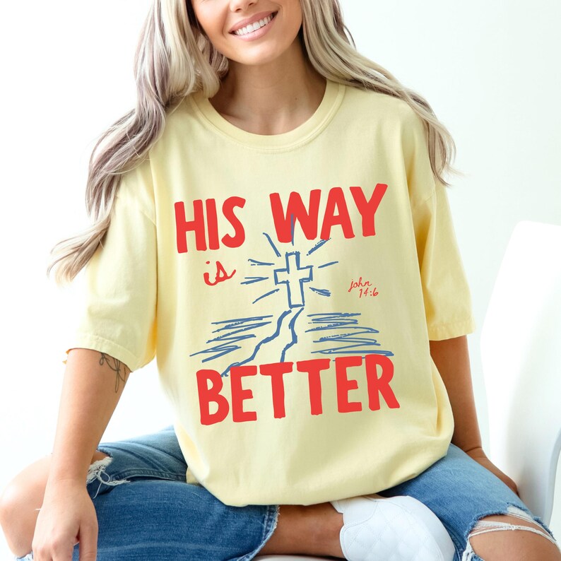 Woman wearing a Christian graphic t-shirt with red text “His Way Is Better” and a blue cross illustration with John 14:6.
