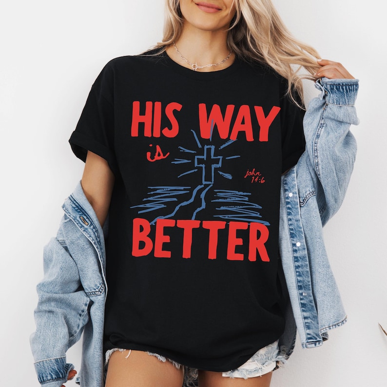 Woman wearing a Christian graphic t-shirt with red text “His Way Is Better” and a blue cross illustration with John 14:6.