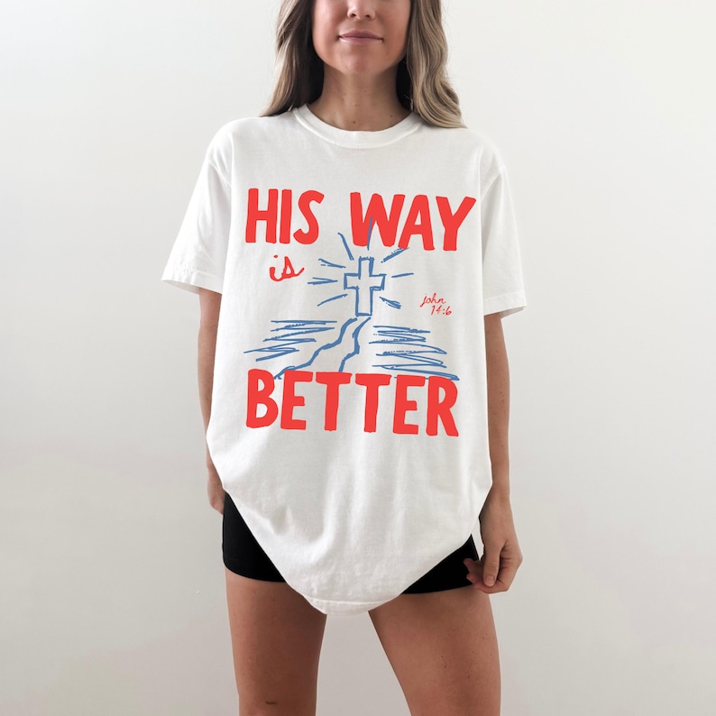 Woman wearing a Christian graphic t-shirt with red text “His Way Is Better” and a blue cross illustration with John 14:6.