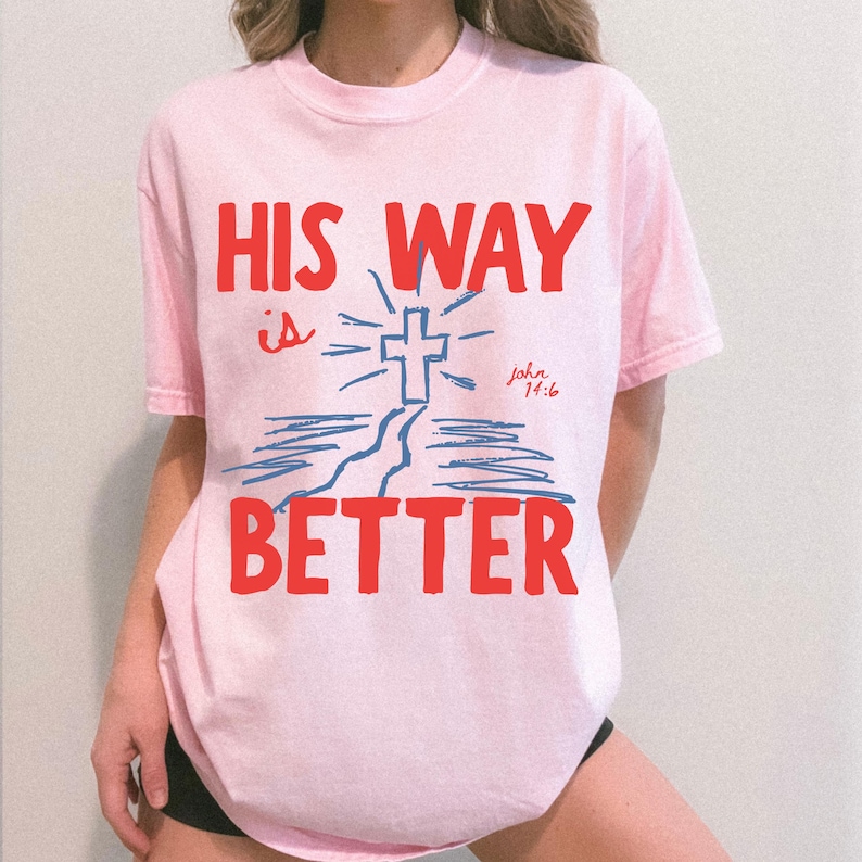 Woman wearing a Christian graphic t-shirt with red text “His Way Is Better” and a blue cross illustration with John 14:6.
