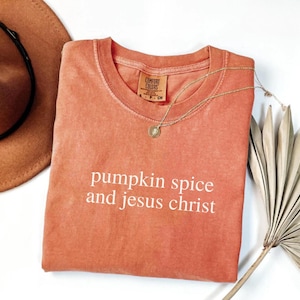 May include: A coral-colored t-shirt with the phrase "pumpkin spice and jesus christ" in white lettering. A gold necklace with a pendant rests on the shirt. A brown hat is visible in the upper left corner.