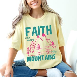 Woman wearing a Christian graphic t-shirt with colorful “Faith can move Mountains” text, pink mountain illustration, and Matthew 17:20 Bible verse.