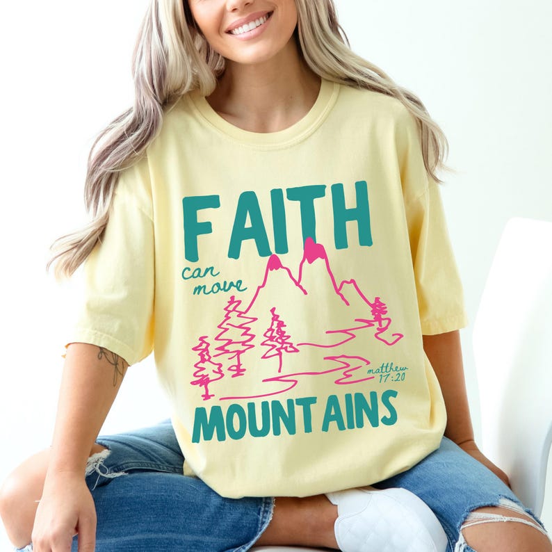 Woman wearing a Christian graphic t-shirt with colorful “Faith can move Mountains” text, pink mountain illustration, and Matthew 17:20 Bible verse.