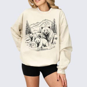 May include: A cream-colored sweatshirt with a black and white illustration of three bears in a forest setting. The bears are standing in a stream with mountains and trees in the background.