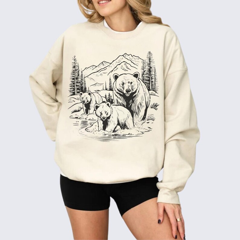 May include: A cream-colored sweatshirt with a black and white illustration of three bears in a forest setting. The bears are standing in a stream with mountains and trees in the background.