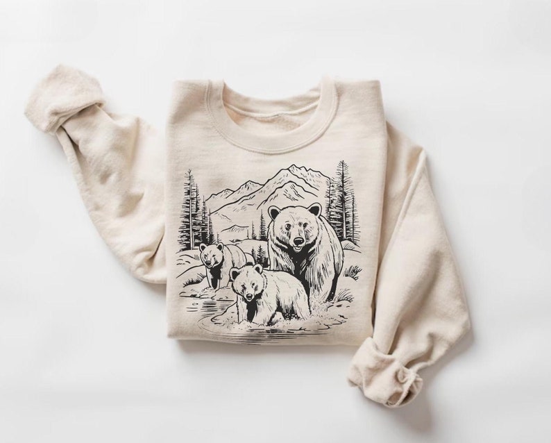 May include: A beige sweatshirt with a black and white illustration of three bears in a mountain landscape. The bears are standing in a stream of water.
