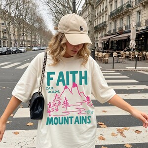 Woman wearing a Christian graphic t-shirt with colorful “Faith can move Mountains” text, pink mountain illustration, and Matthew 17:20 Bible verse.