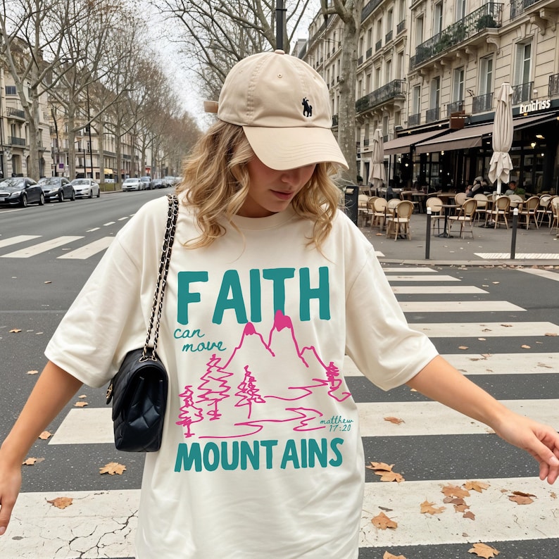 Woman wearing a Christian graphic t-shirt with colorful “Faith can move Mountains” text, pink mountain illustration, and Matthew 17:20 Bible verse.