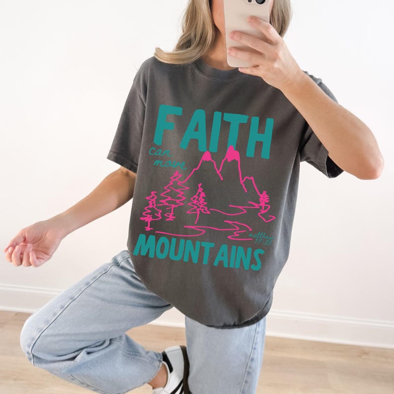 Woman wearing a Christian graphic t-shirt with colorful “Faith can move Mountains” text, pink mountain illustration, and Matthew 17:20 Bible verse.