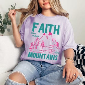Woman wearing a Christian graphic t-shirt with colorful “Faith can move Mountains” text, pink mountain illustration, and Matthew 17:20 Bible verse.
