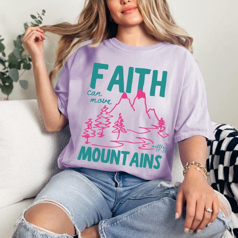 Woman wearing a Christian graphic t-shirt with colorful “Faith can move Mountains” text, pink mountain illustration, and Matthew 17:20 Bible verse.