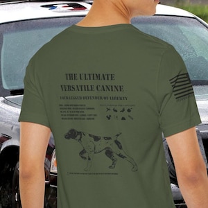 May include: A man wearing a green t-shirt with a black and white illustration of a dog on the back. The shirt says "The Ultimate Versatile Canine" and "The Unlikely Defender of Liberty". The man is standing in front of a black and white police car.