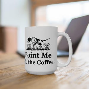 May include: White ceramic coffee mug with a black and white illustration of a dog in tall grass. The text "Point Me To the Coffee" is printed below the dog illustration. The mug has a large handle.
