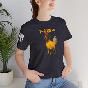 May include: Navy blue t-shirt with the text "F-CAW-F" and a dog dressed as a rooster. The dog has a yellow body, a red wattle, and a brown head. The shirt also has a small American flag on the left sleeve.