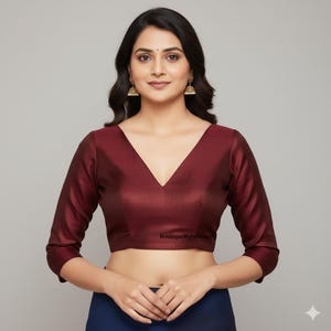 Puede incluir: Una blusa burdeos con cuello en V profundo y mangas tres cuartos. La blusa tiene un brillo sutil y un corte entallado. El texto "BoutiquebyAnshary" está impreso en la parte inferior de la blusa. La blusa se combina con una prenda azul marino.