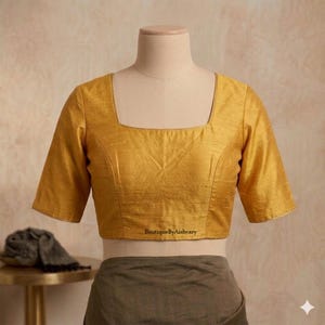 Puede incluir: Blusa amarillo mostaza con cuello cuadrado y mangas cortas. La blusa se exhibe en un maniquí. El texto "BoutiqueByAishvary" está impreso en la parte inferior de la blusa.