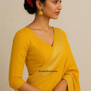 Könnte beinhalten: Eine Frau in einem senfgelben Sari und einer passenden Bluse. Die Bluse hat einen V-Ausschnitt und Dreiviertelärmel. Goldene Ohrringe und eine rote Blume im Haar sind zu sehen. Der Text "BoutiqueByAishvary" ist sichtbar.